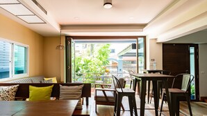 A modern dining area with a wooden table and chairs, a sofa with cushions, and a view of greenery through large windows. - Maneeya Park Residence (Bangkok)
