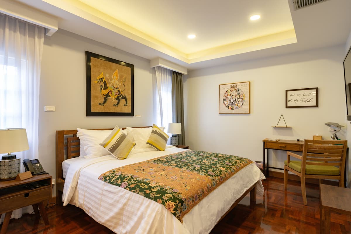 A hotel room with a bed, bedside table with a lamp, a desk with a chair, and artwork on the walls.