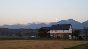 A house with a dark roof and white walls, surrounded by a field and mountains in the background. - Gufo no Mori Kamifurano (Kamifurano)