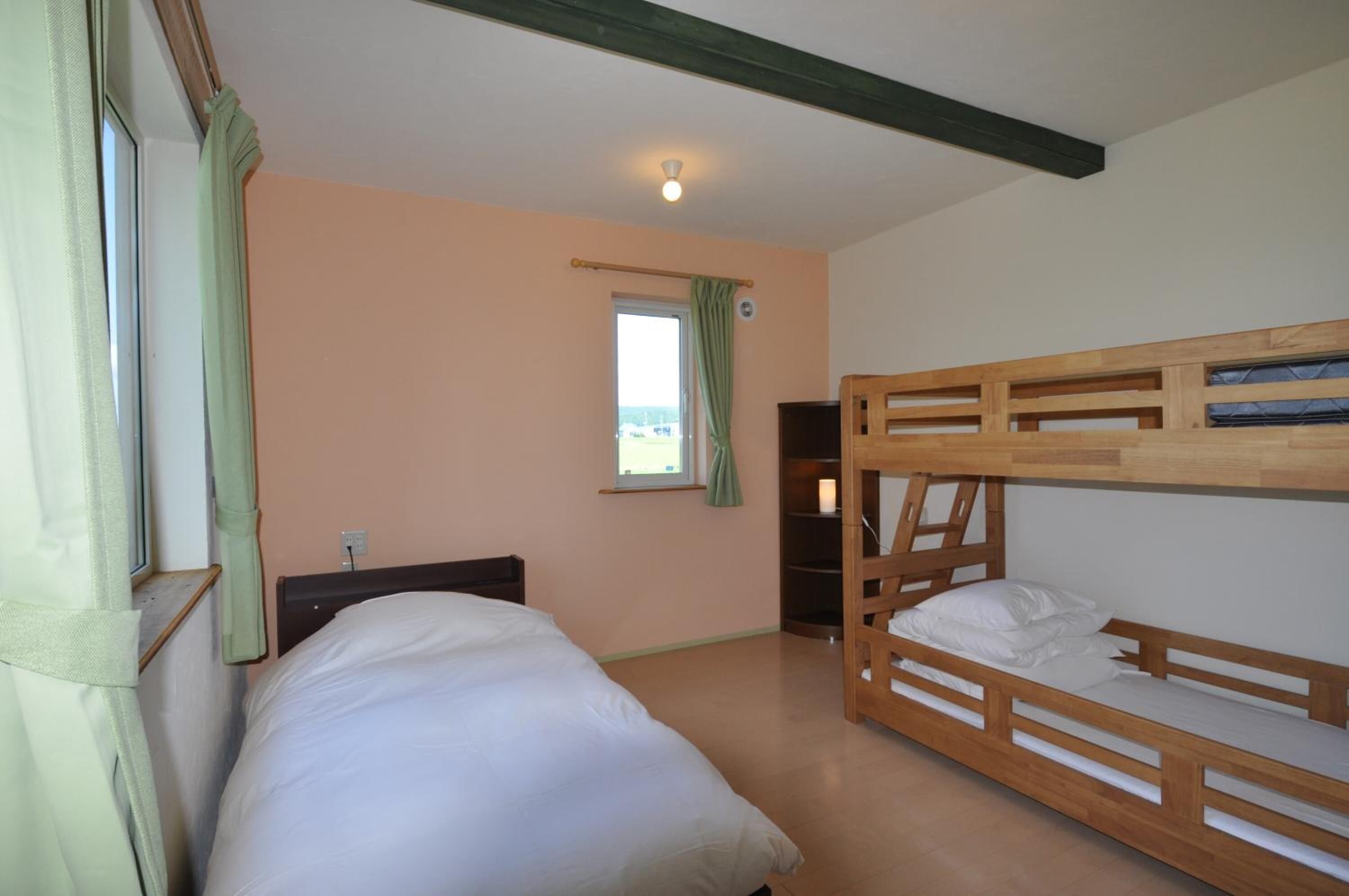 A room with a bunk bed, a single bed, a window with curtains, and a small desk with a lamp.
