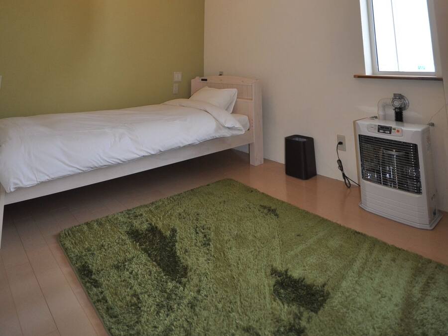 Habitación pequeña con cama individual, alfombra verde, vigas de madera, ventana y un calentador.