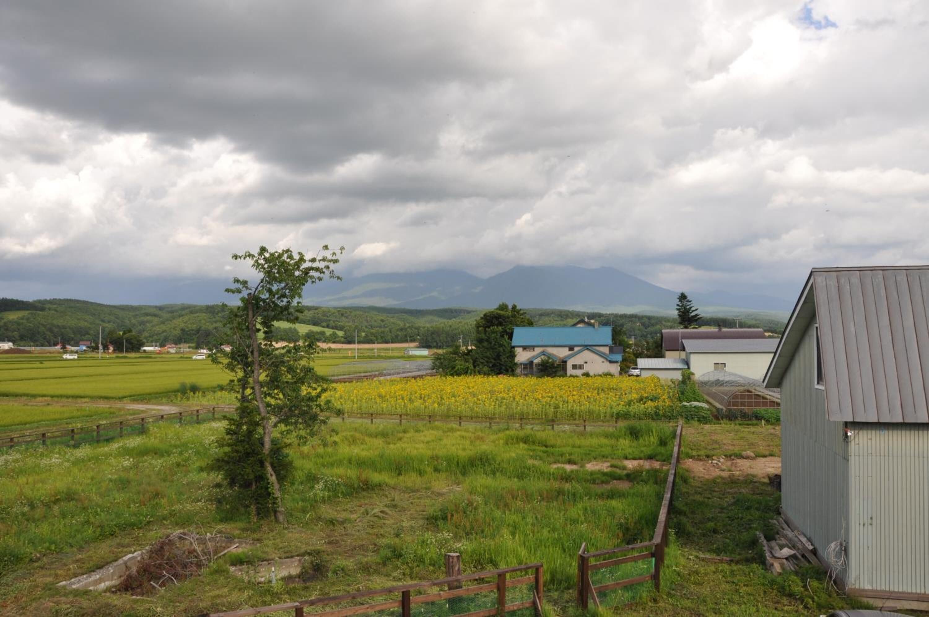 A rural landscape with fields, a small building, and distant mountains under a cloudy sky.