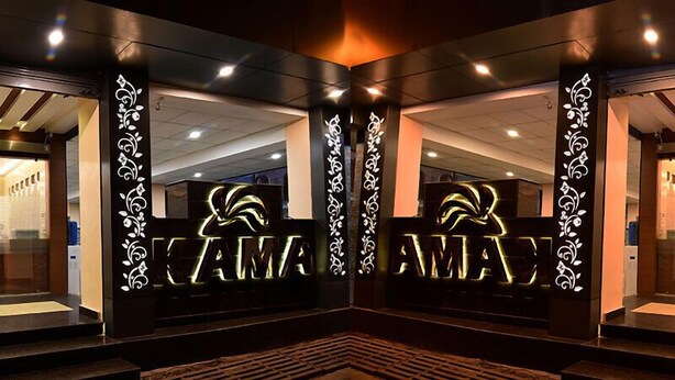 Een hotelingang met verlicht 'KAMA' uithangbord, een metselstenen vloer en groene zitgedeeltes.