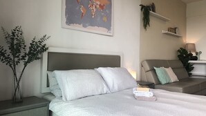 A neatly arranged bedroom with a bed, a nightstand, a world map on the wall, a sofa, and a small table with a lamp and decorative items.