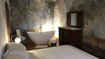 Une chambre dotée d’une grande baignoire, d’une commode en bois, d’un lit avec du linge de maison blanc et d’un mur sobre agrémenté d’un miroir.