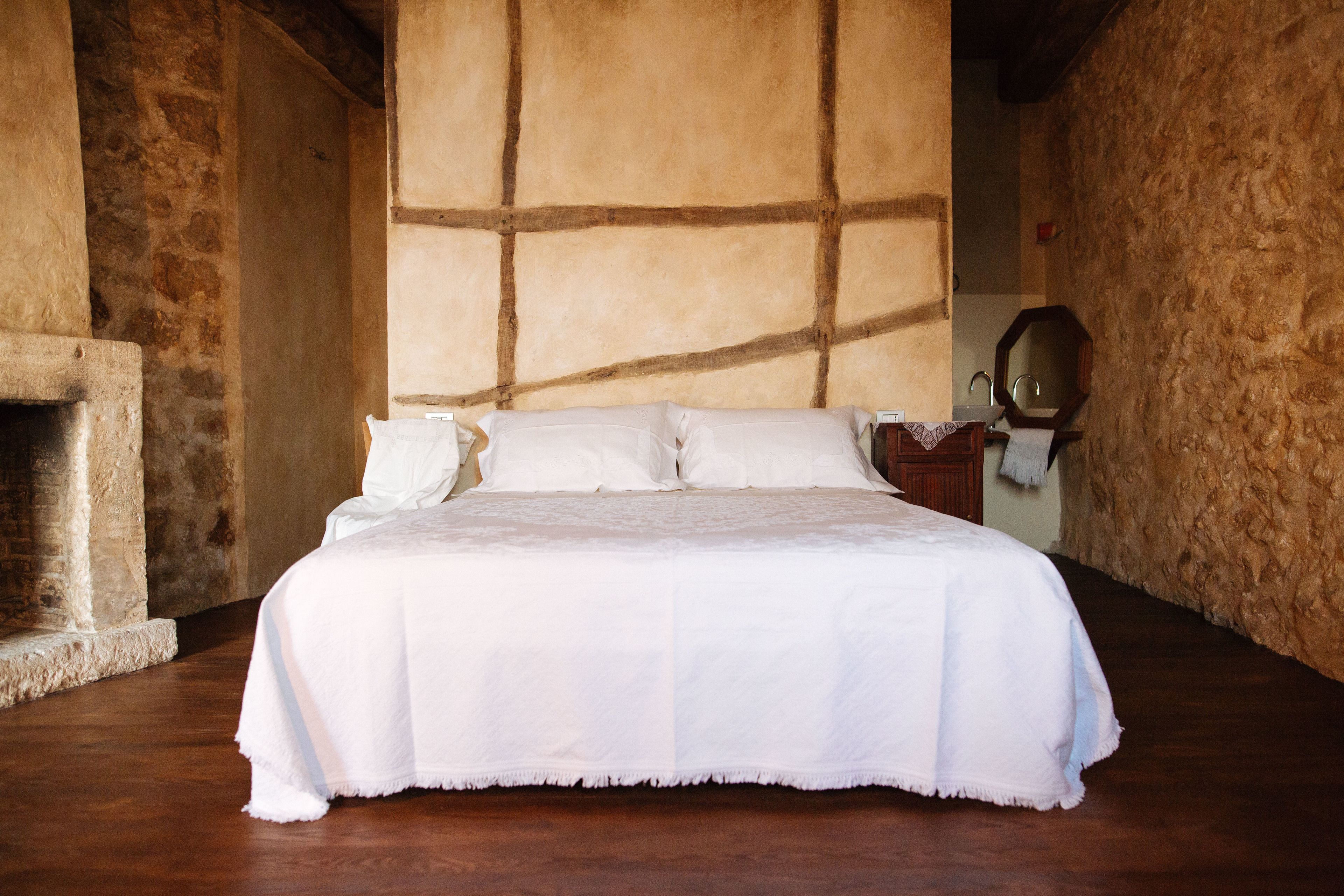 Un grand lit recouvert de draps blancs, dans une chambre rustique aux murs de pierre et dotée d’une cheminée.