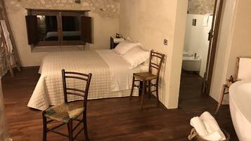 A bedroom with a large bed, two wooden chairs, a stone wall, and a view of a bathroom.