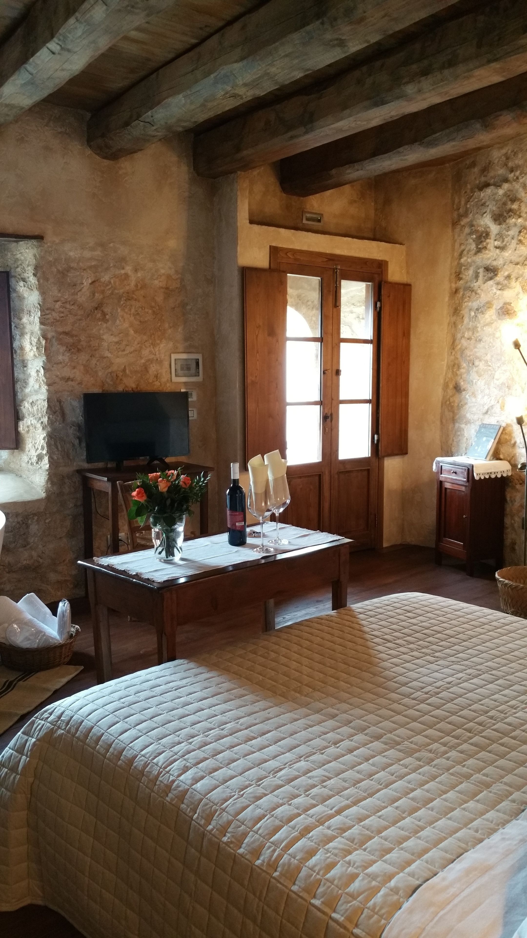A rustic room with a bed, a wooden table with a vase of flowers, a bottle of wine, and a television on a wooden stand.