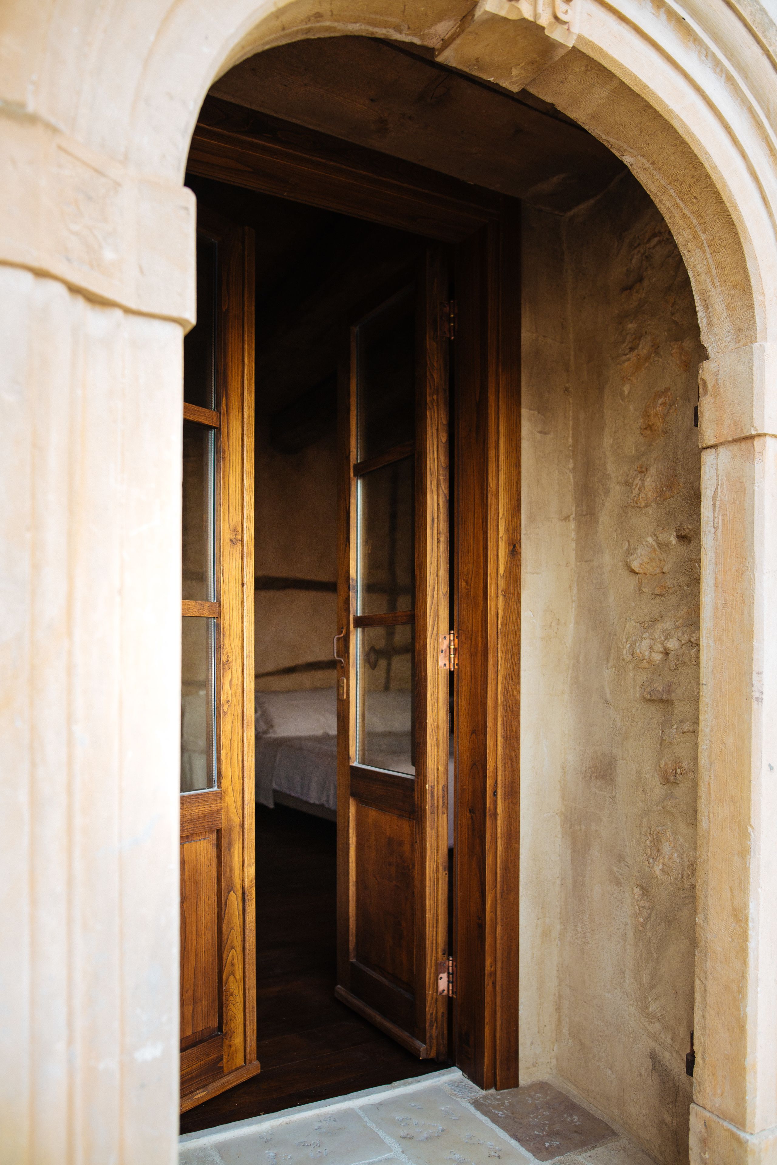 Uma porta em arco dá acesso a um quarto com cama e molduras de porta de madeira.