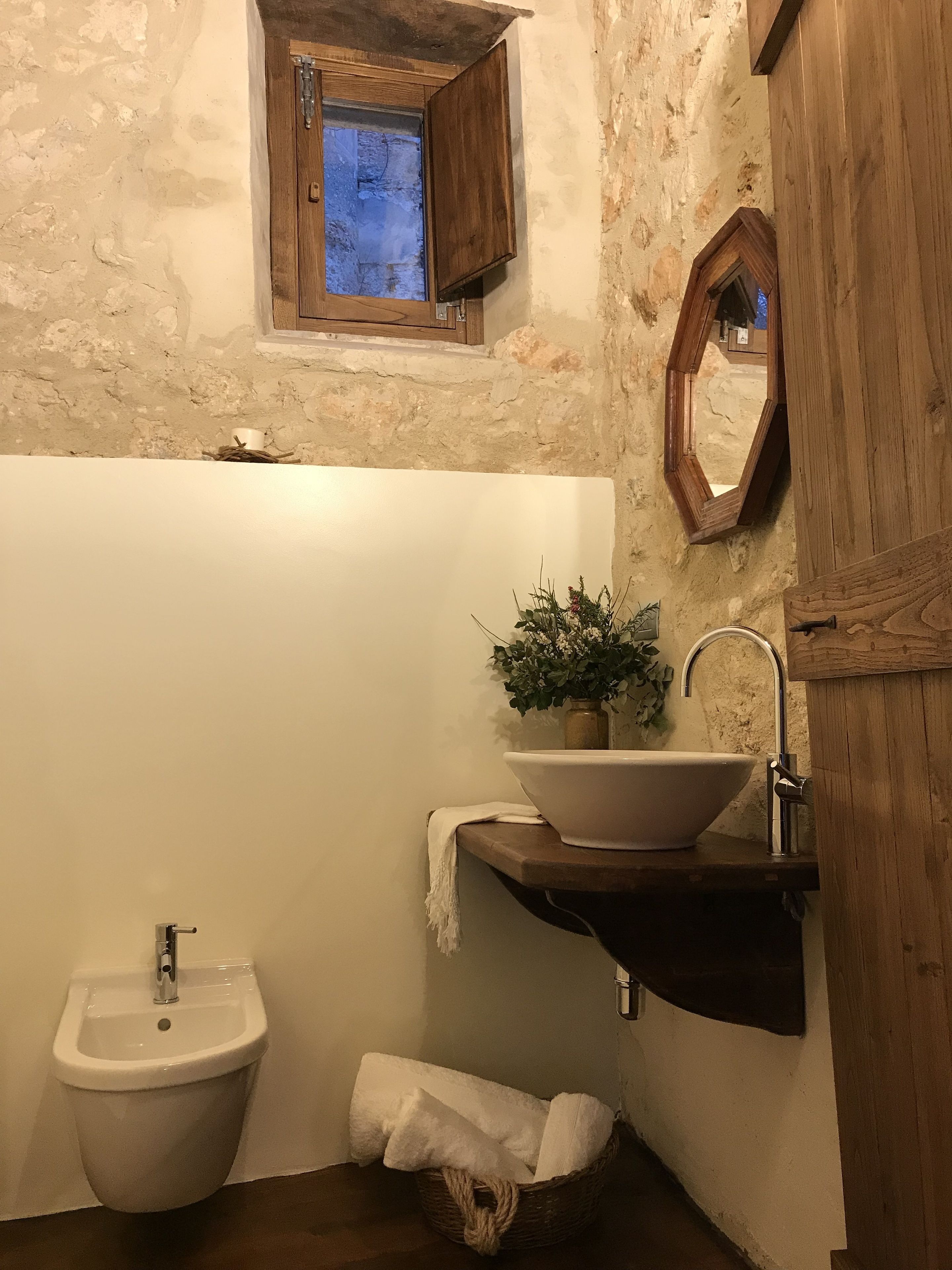 A rustic bathroom with a wooden cabinet, a small window, a sink with a plant, and a bidet.