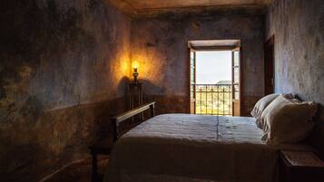 Ein schwach beleuchtetes Zimmer mit einem großen Bett, einem kleinen Tisch mit einer Lampe und Blick durch eine geöffnete Tür.