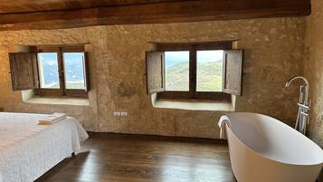 A rustic bedroom with wooden floors, a large bathtub, and two windows with wooden shutters.