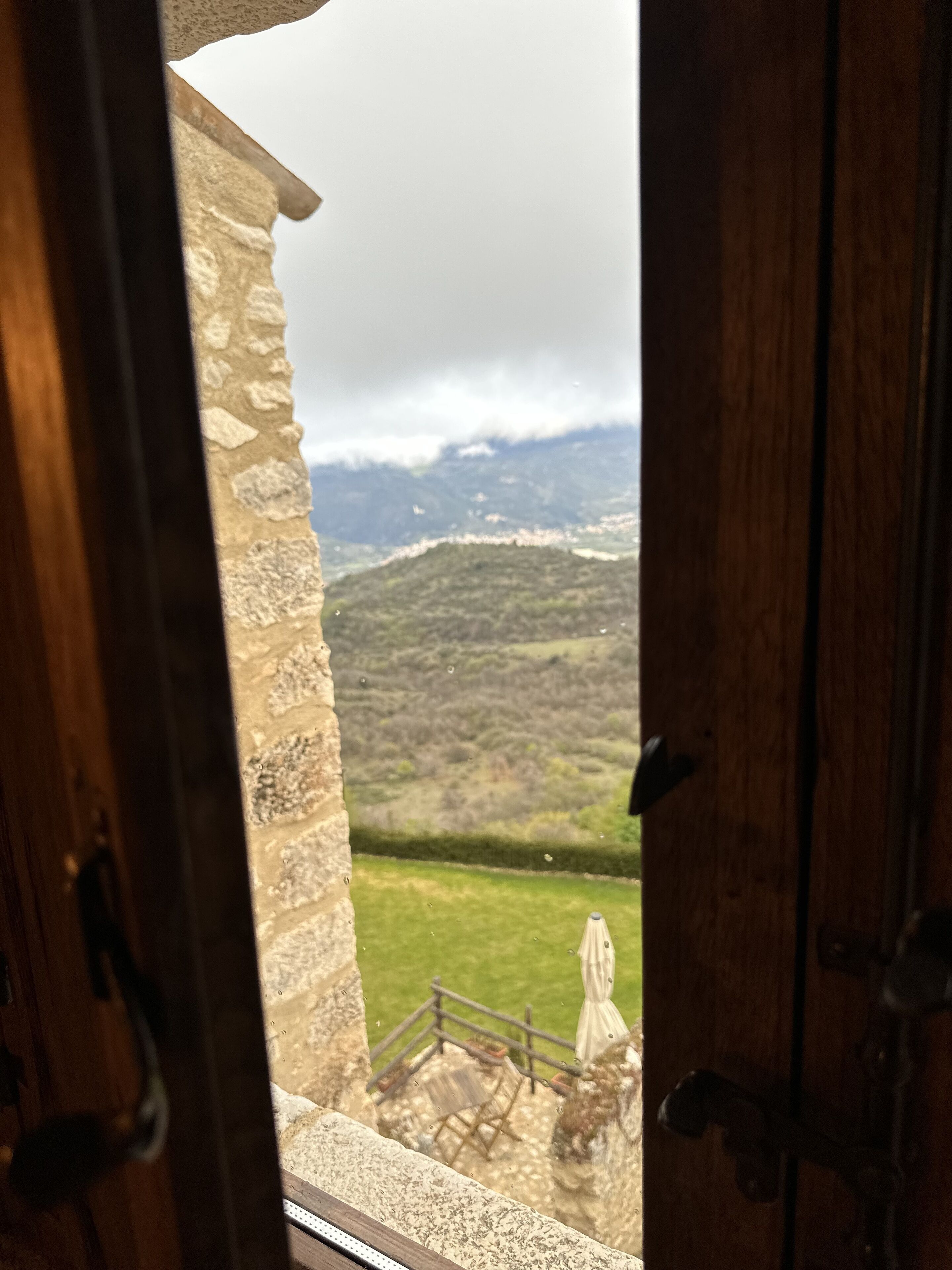 Blick durch eine offene Tür auf einen Balkon mit einer Steinsäule, einer Rasenfläche und einer Berglandschaft im Hintergrund.