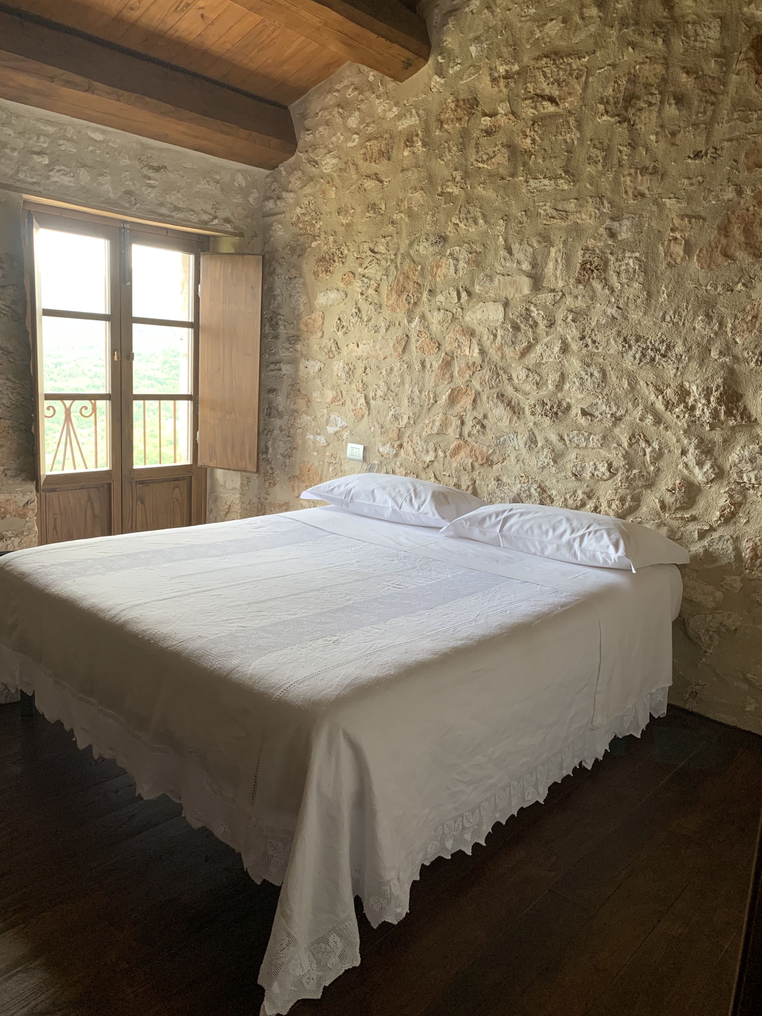 A bedroom with a stone wall, a wooden ceiling, and a large bed with white linens.