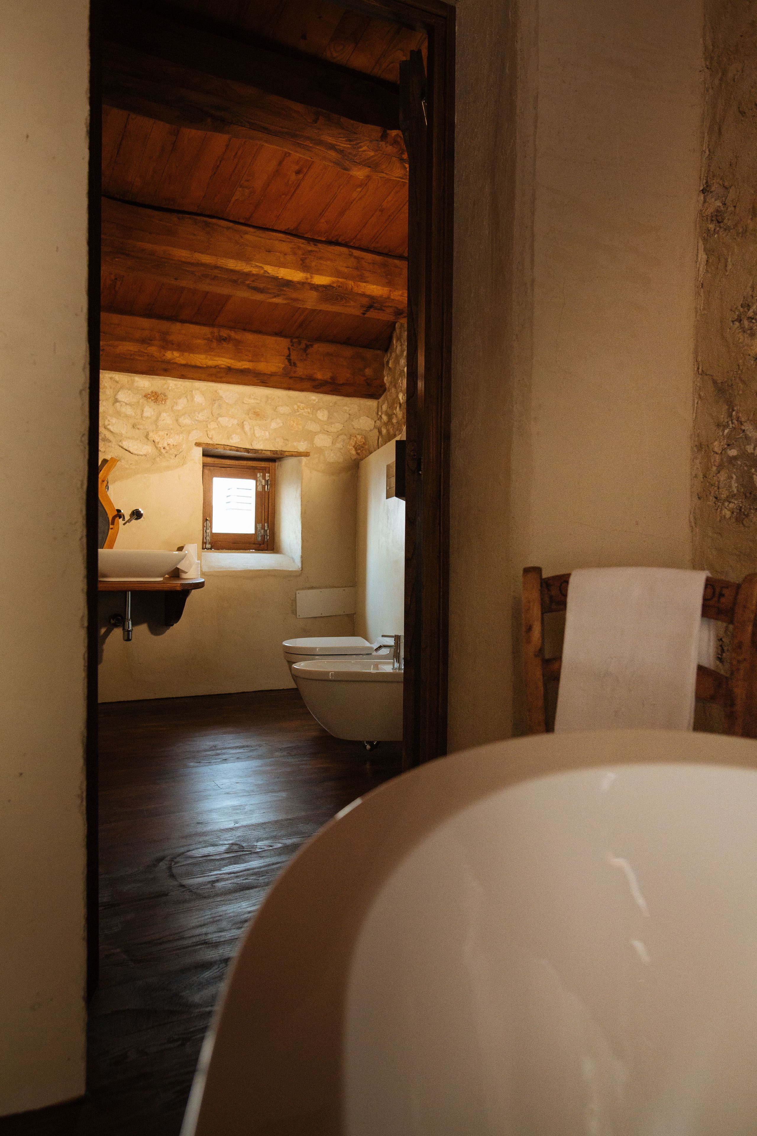 A rustic bathroom with a wooden ceiling, a stone wall, a wooden chair, and a bathtub.