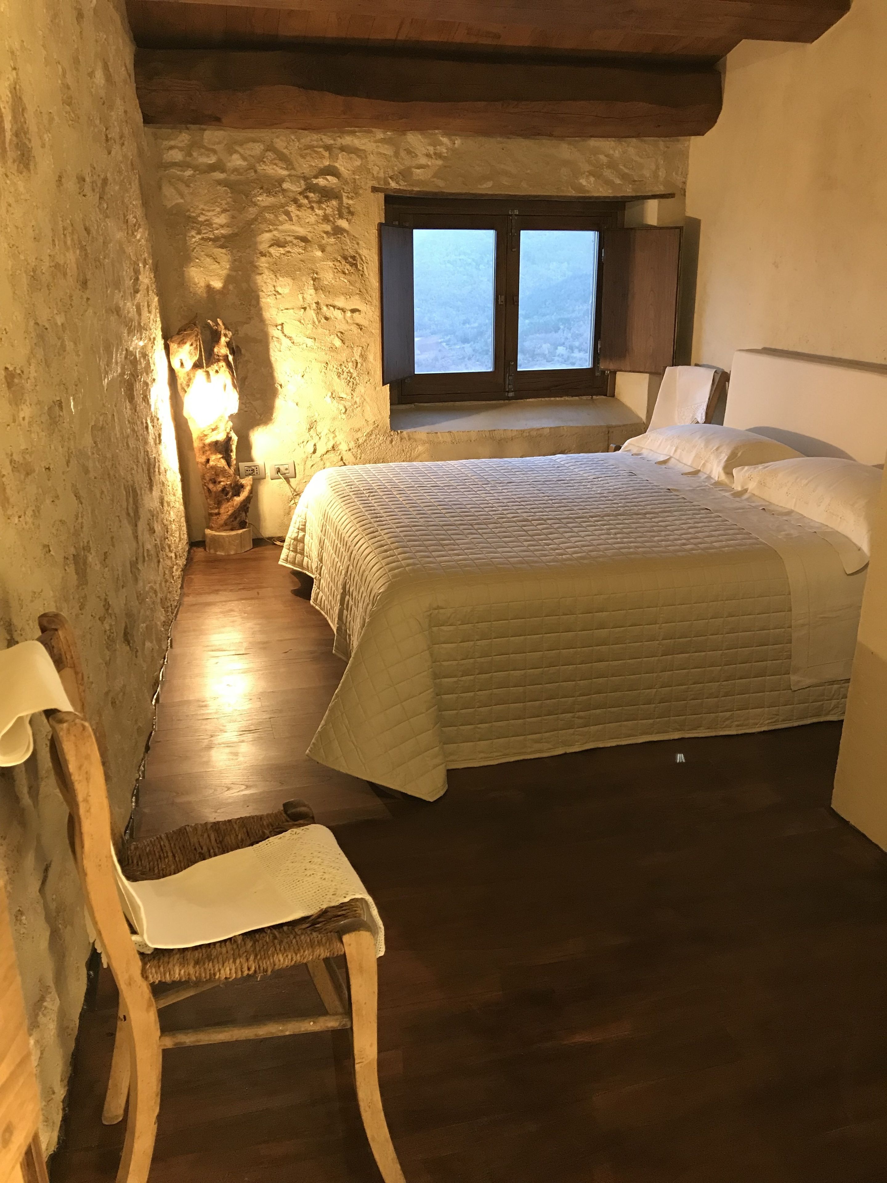 A cozy bedroom with a wooden bed, a chair, a nightstand, and a window with a view.
