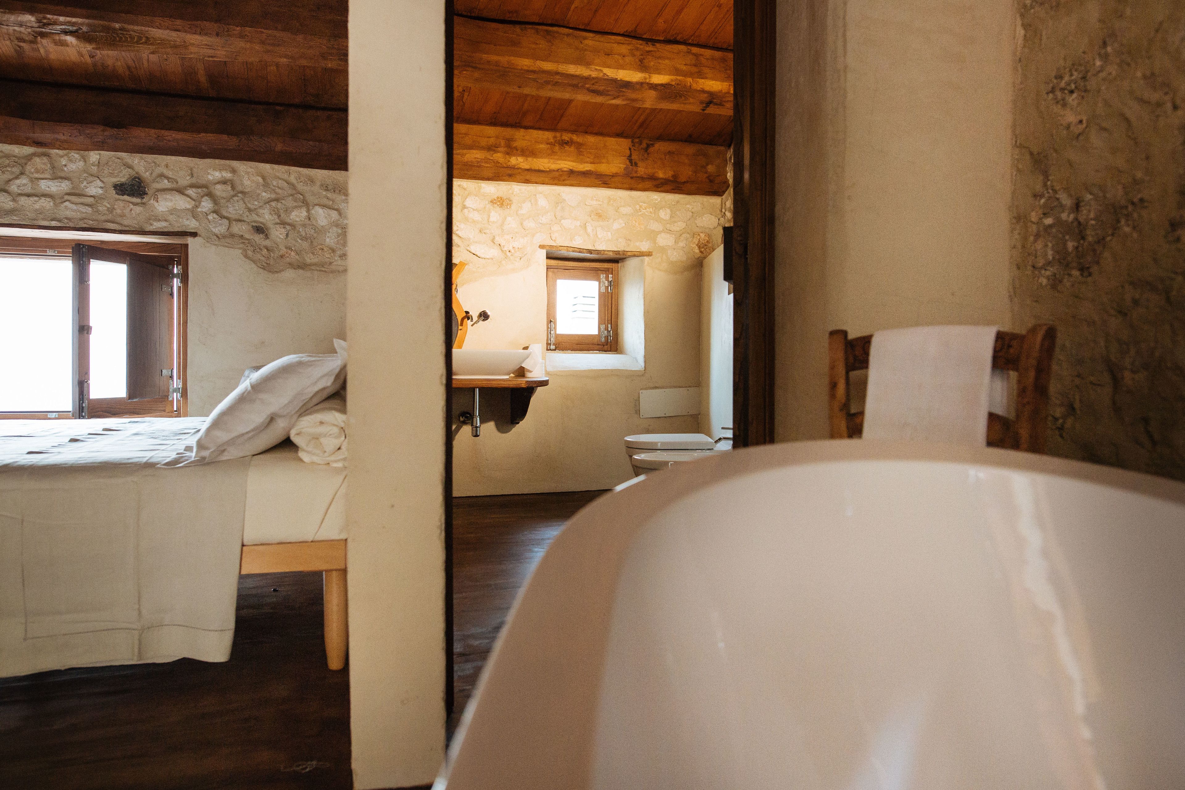 A rustic room with a wooden ceiling, a stone wall, a bed with a white bedspread, a wooden chair, and a bathtub.