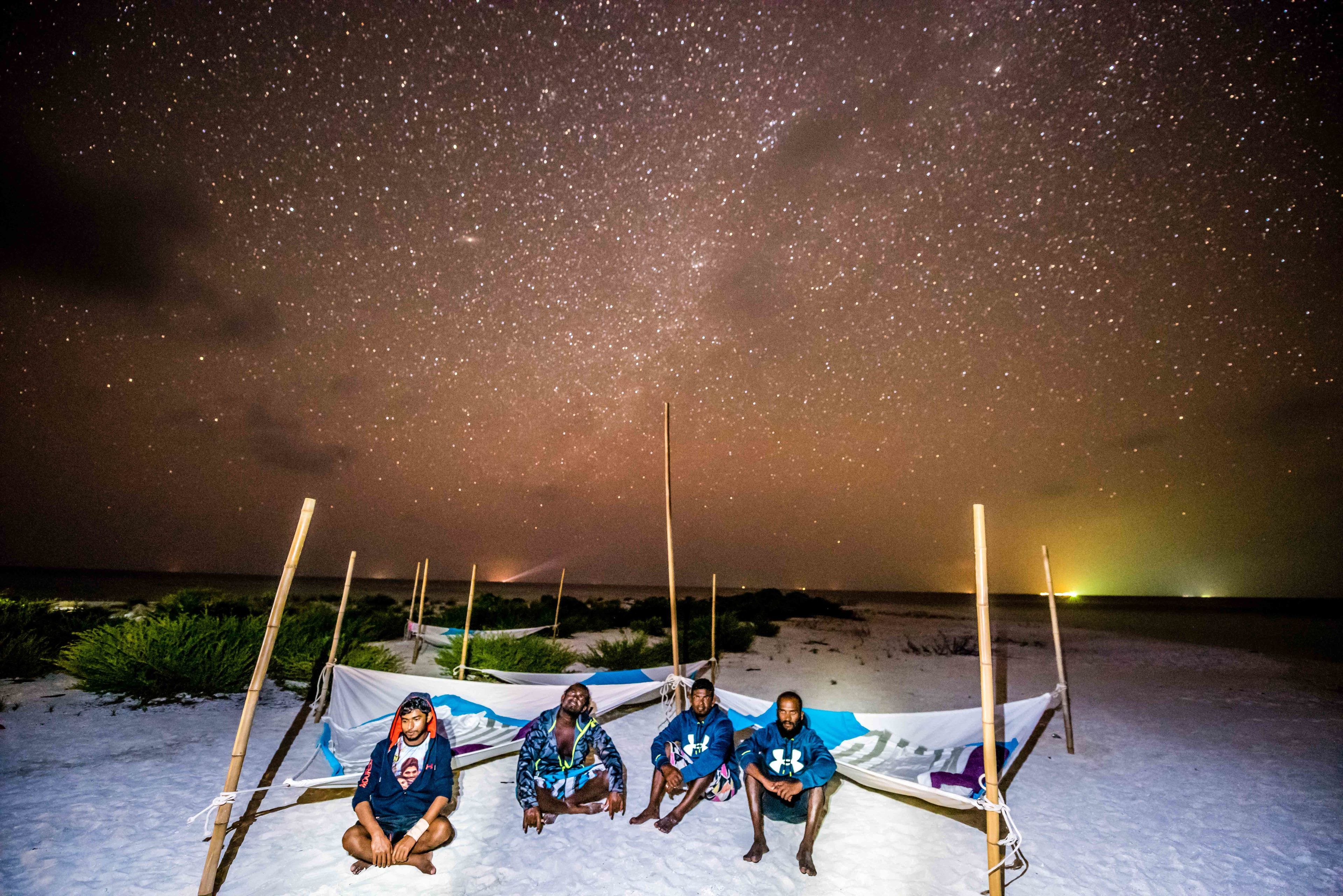 Three people sitting on a beach under a starry sky with a tent in the background.