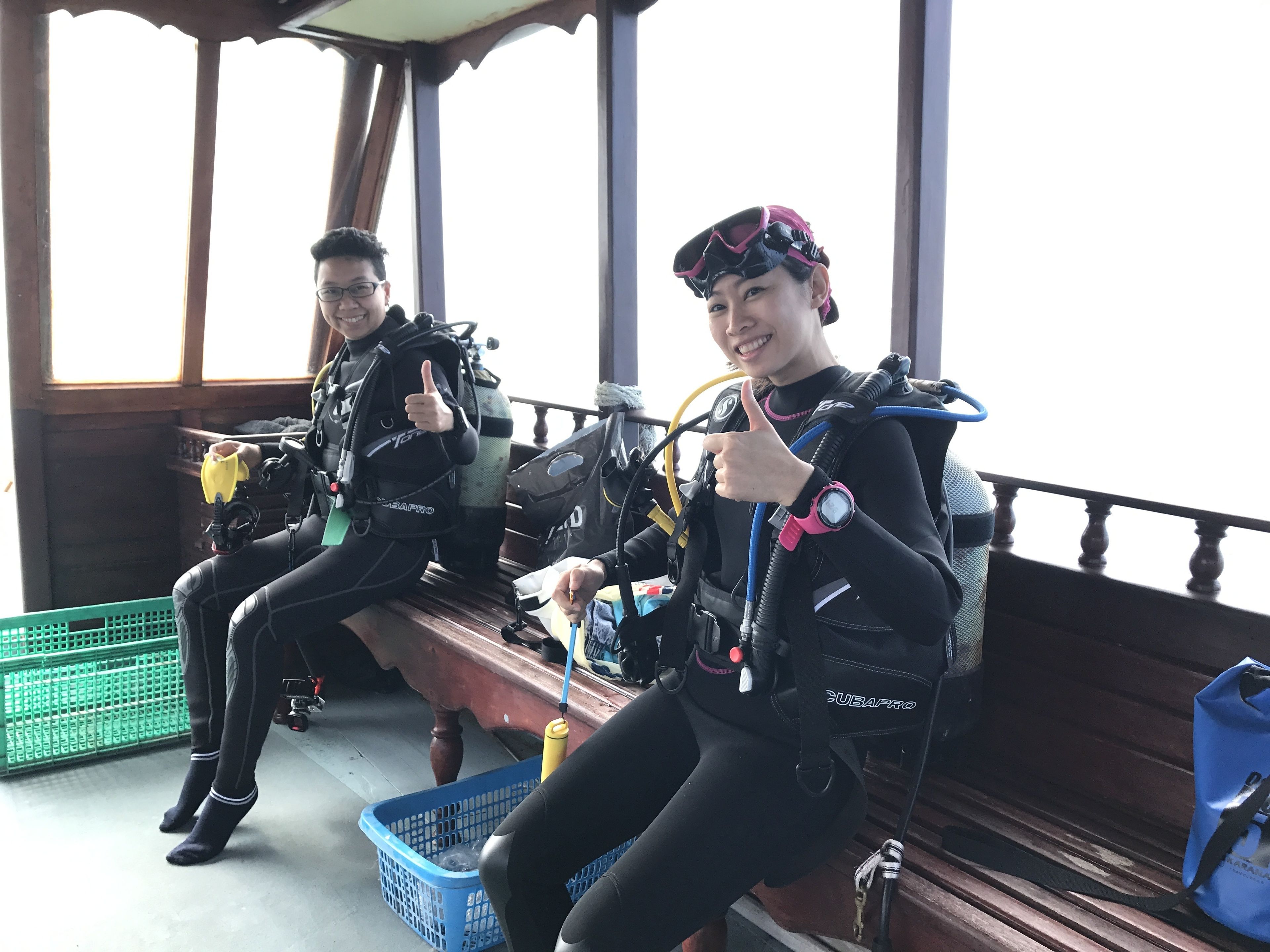 Dos personas con equipo de buceo sentadas en un banco de madera, cerca de una cesta verde y una bolsa azul.