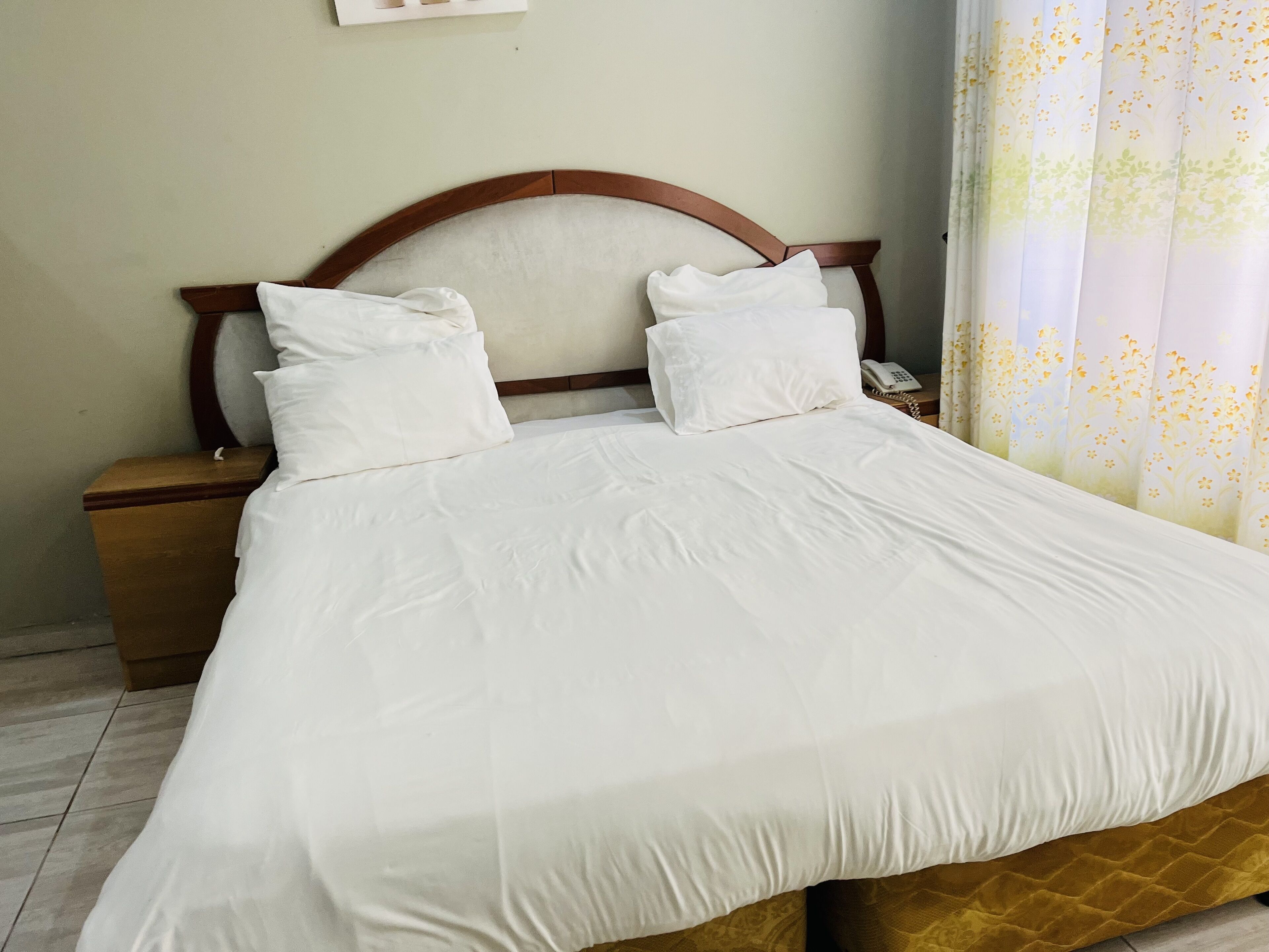 A neatly made bed with white linens and a wooden headboard, a wooden nightstand with a telephone, and a floral curtain.
