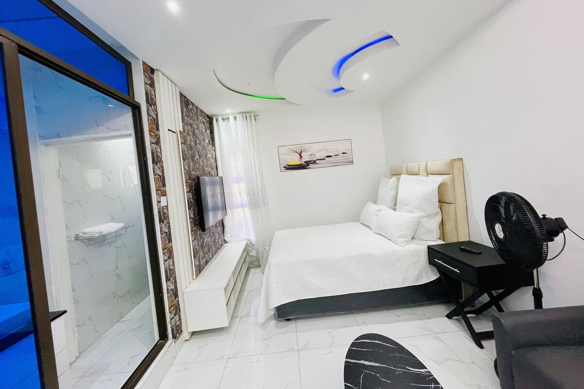 A modern hotel room with a bed, a desk, a chair, a fan, and a TV.