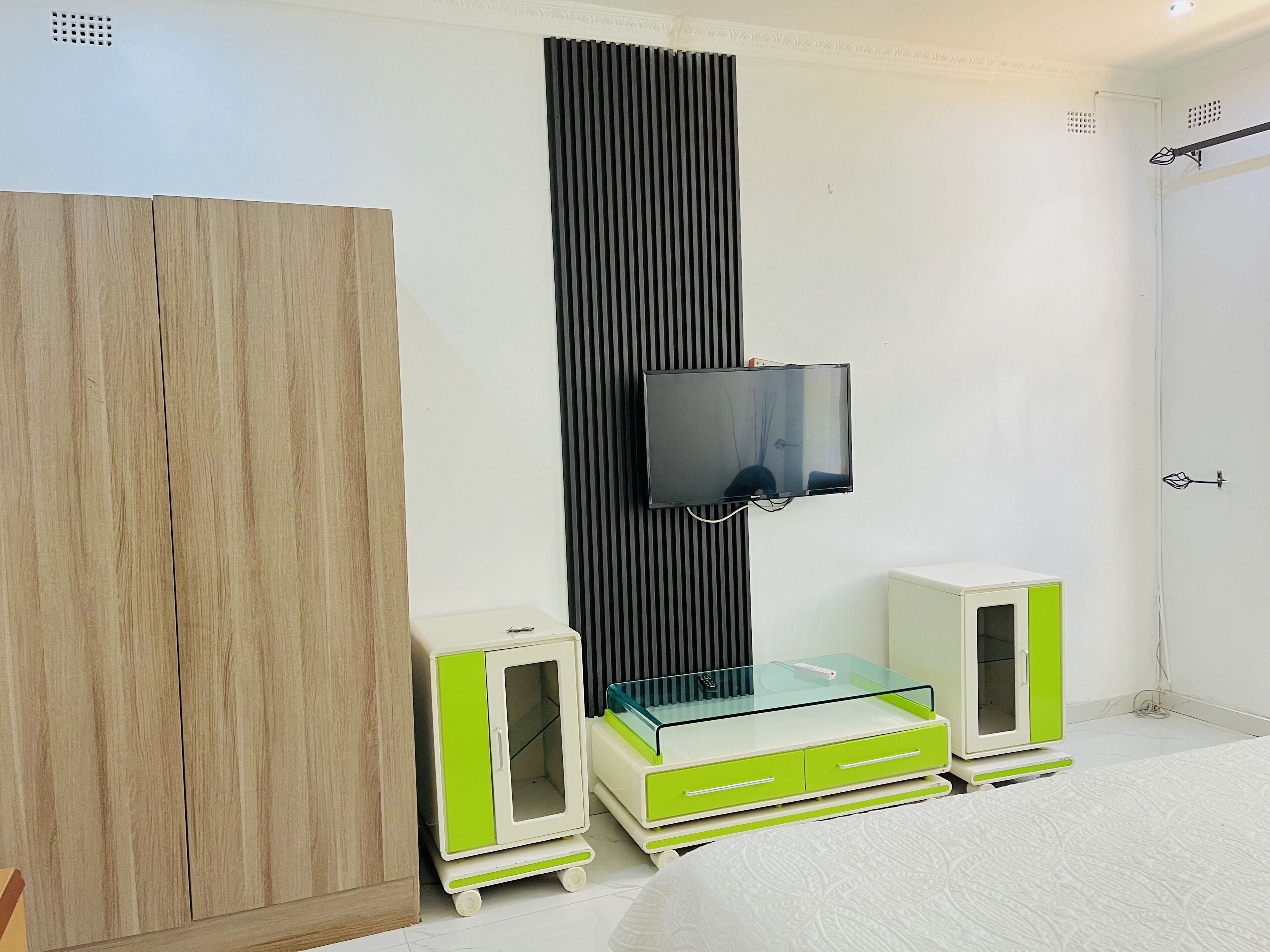 A bedroom with a wooden wardrobe, a black vertical panel, a mounted TV, and a green and white bedside table with drawers.