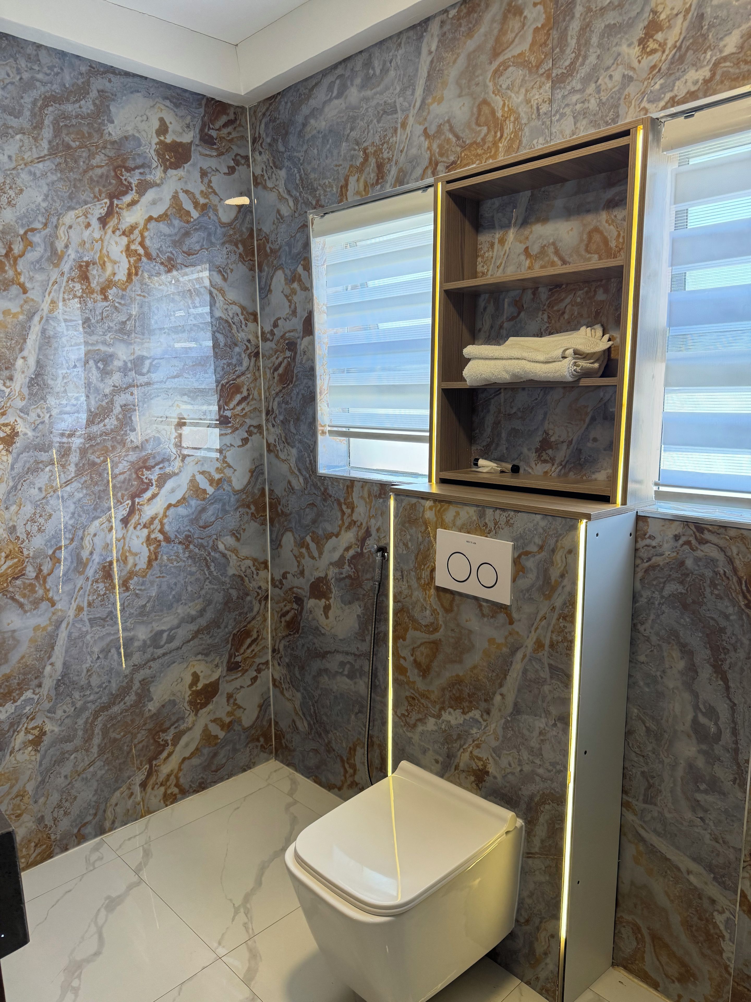 Ein Badezimmer mit Marmorwand, Toilette, Ablagefläche mit Handtüchern und Fenster mit Jalousien.