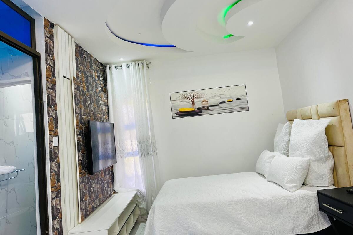 A bedroom with a bed, a TV mounted on the wall, a painting, and a ceiling with recessed lighting.