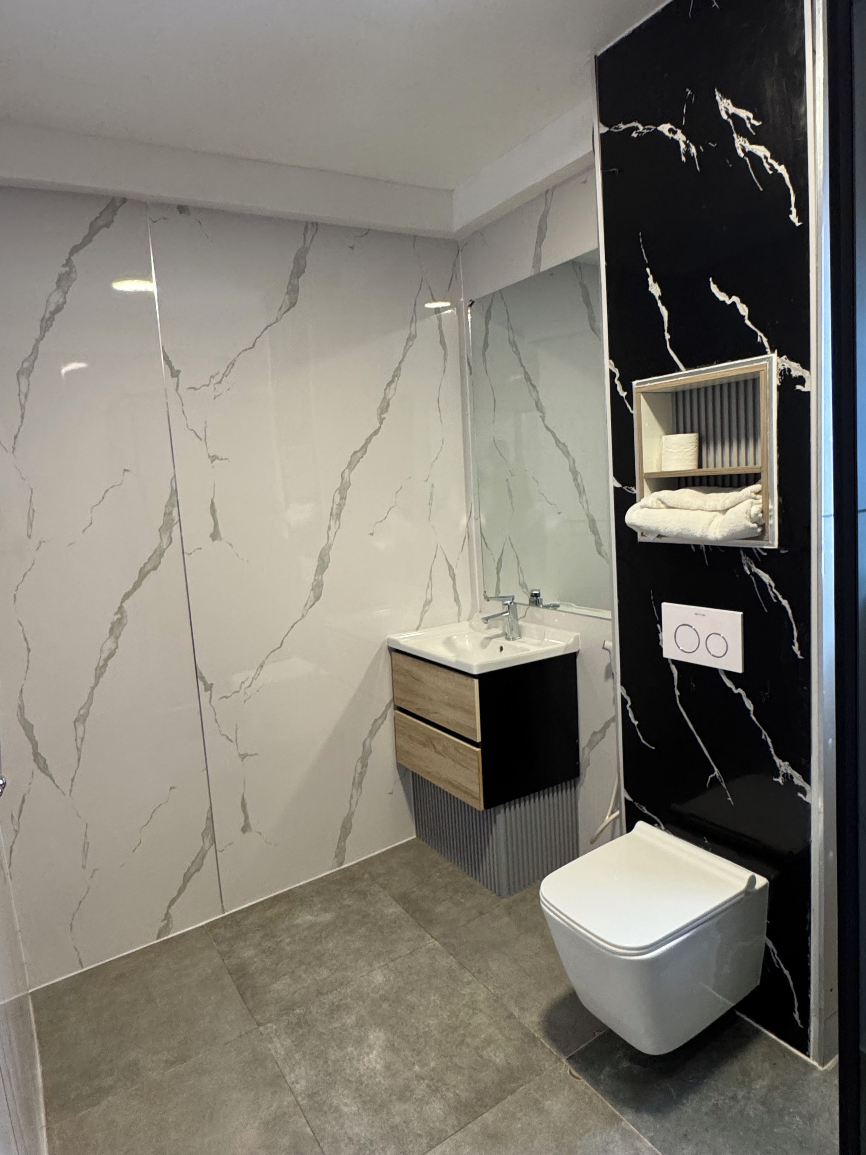 A modern bathroom with a marble wall, a white toilet, a vanity with a mirror, and a towel rack.