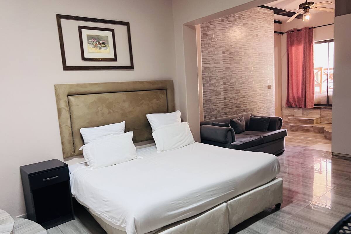 A modern hotel room with a large bed, a headboard, a small bedside table, a sofa, and a view of a balcony.