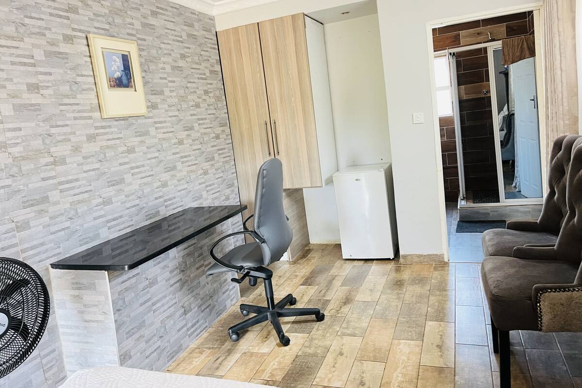 A modern living room with a grey tiled wall, a black office chair, a white refrigerator, and a brown sofa.