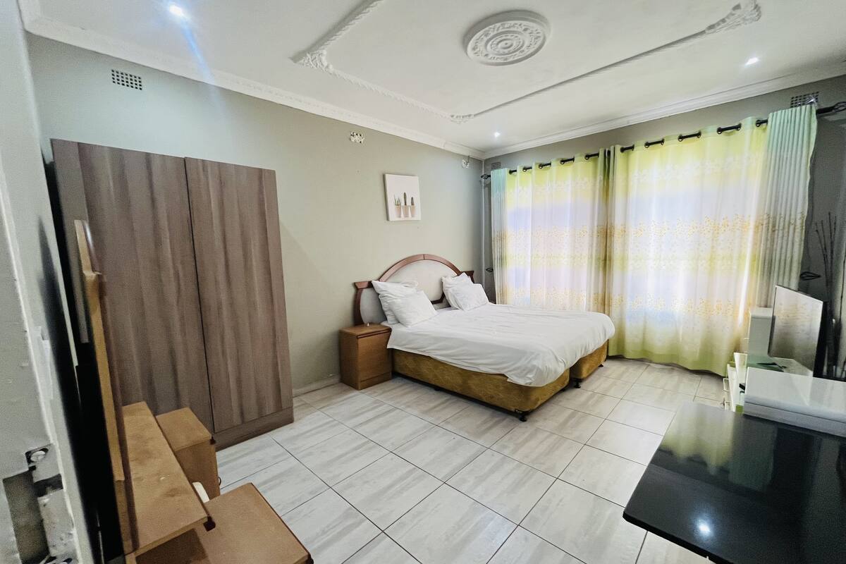 A hotel room with a bed, a nightstand, a wooden wardrobe, a mirror, and a desk with a chair.