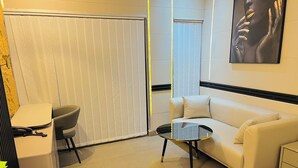 A modern hotel room with a white sofa, a small round table, a chair, and a large window with blinds. - Highlands Lodges and Apartments (Harare)