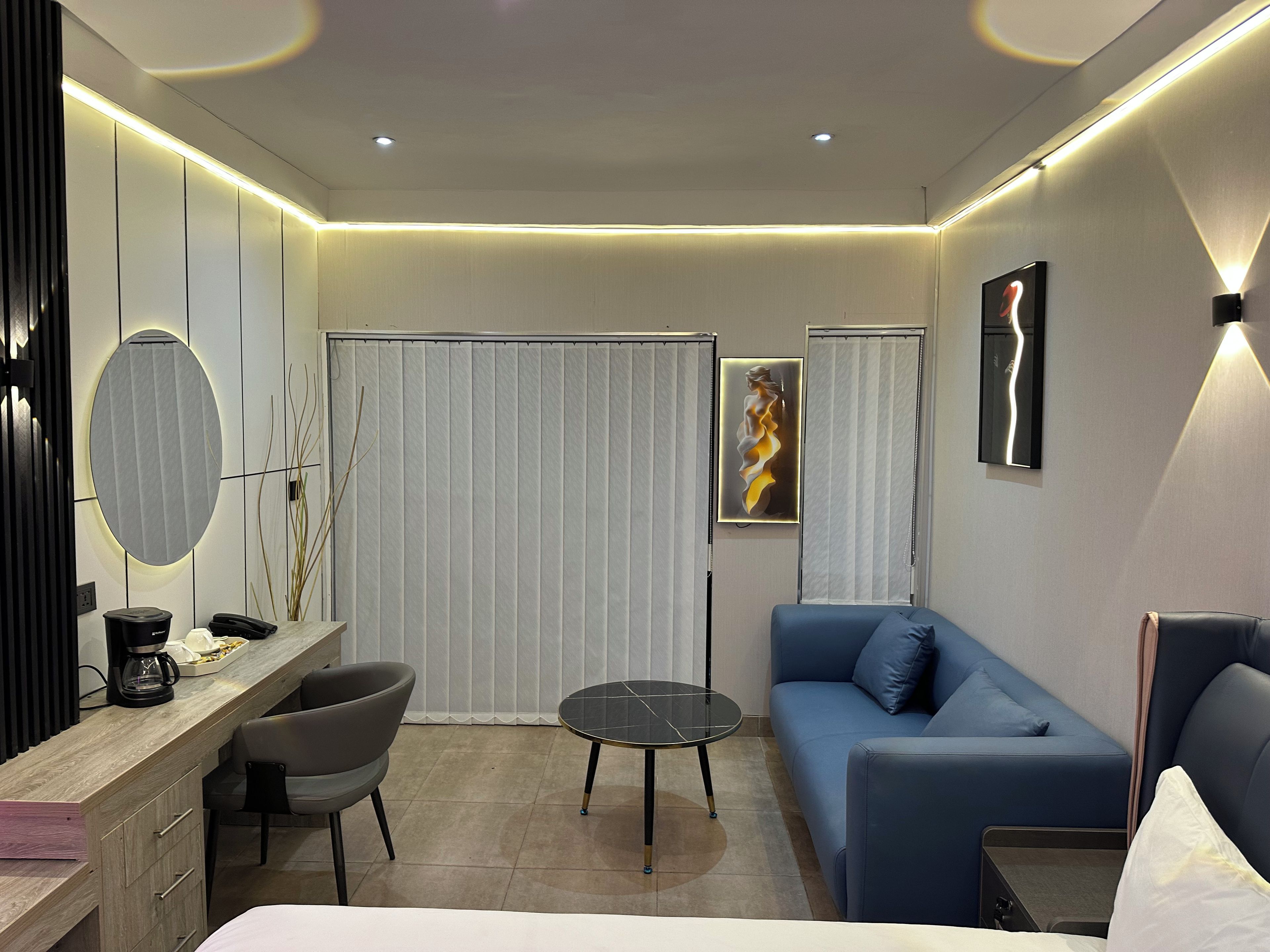 A modern hotel room with a bed, a desk with a chair, a small round table, a sofa, and a large mirror.