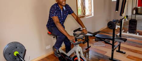 A person using an exercise bike in a room with wooden flooring, a window with bars, and various fitness equipment.