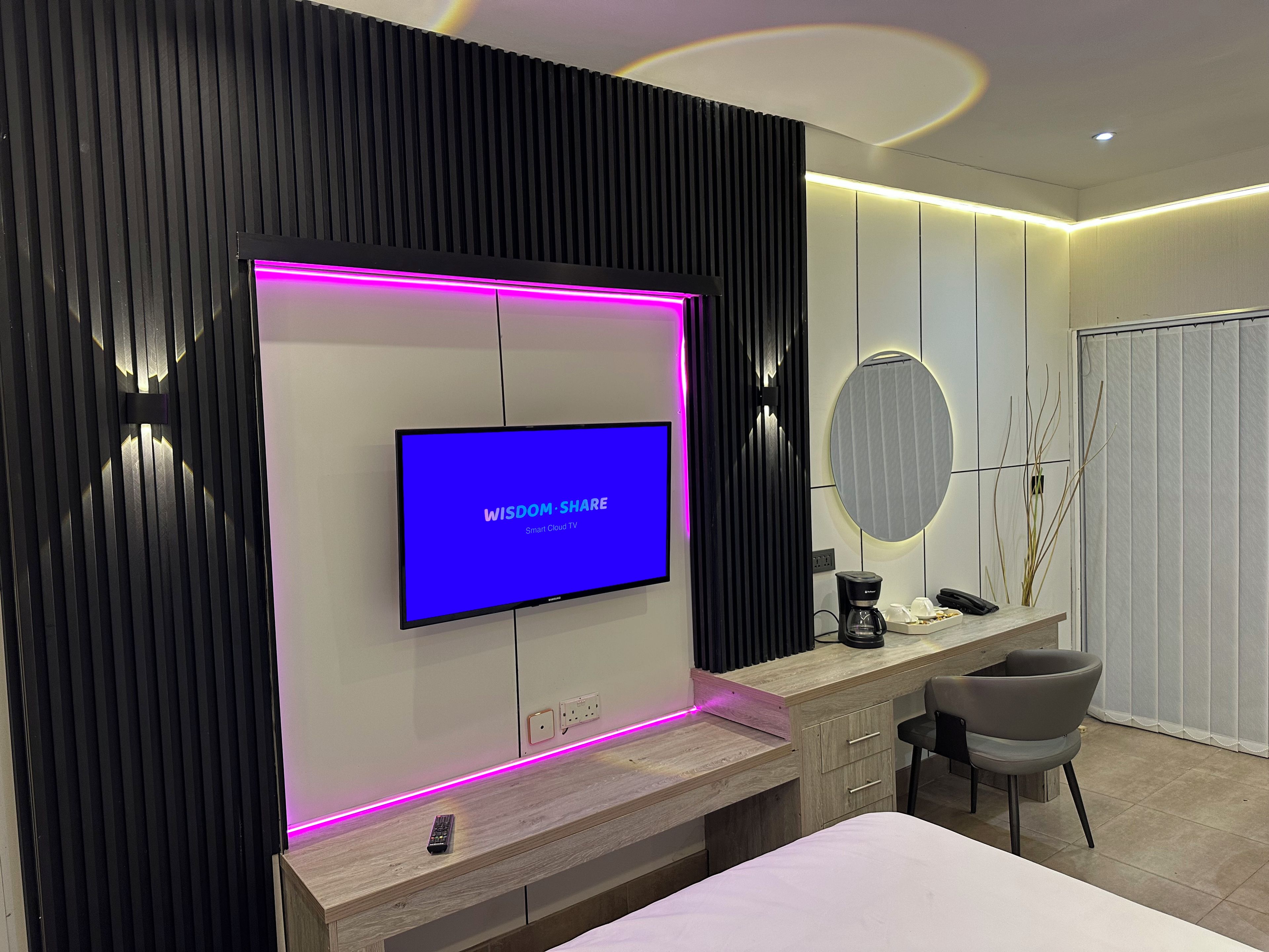 A modern hotel room with a flat-screen TV, a wooden desk, a chair, and a bedside table.