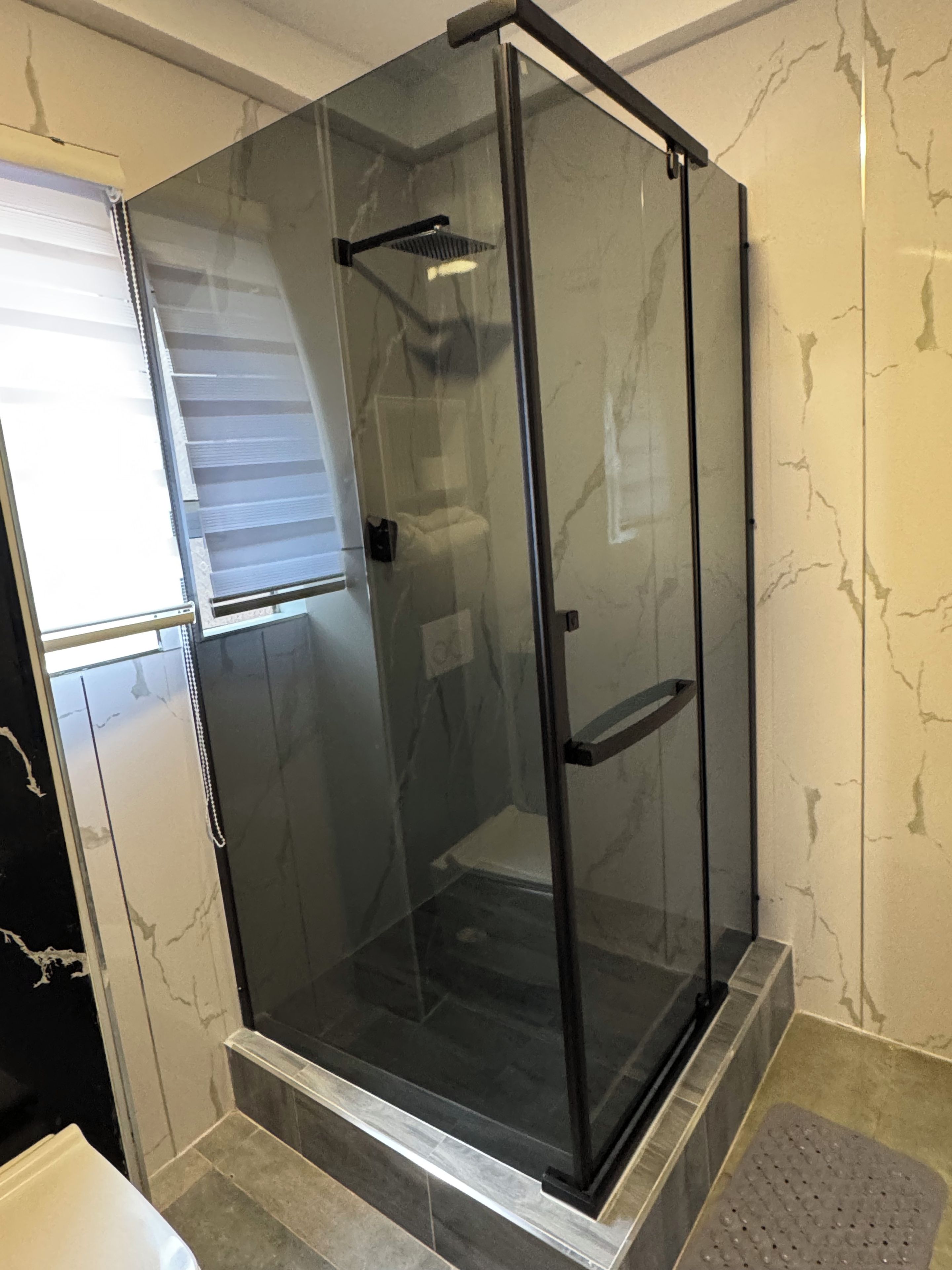 Cabina de ducha con paredes de cristal, marco negro, puerta blanca y piso de baldosas.