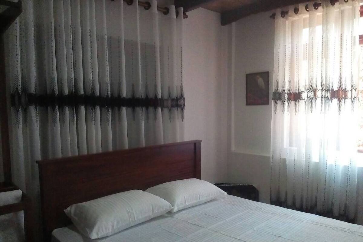 A bedroom with a wooden bed, white bedding, and sheer curtains with a decorative pattern.
