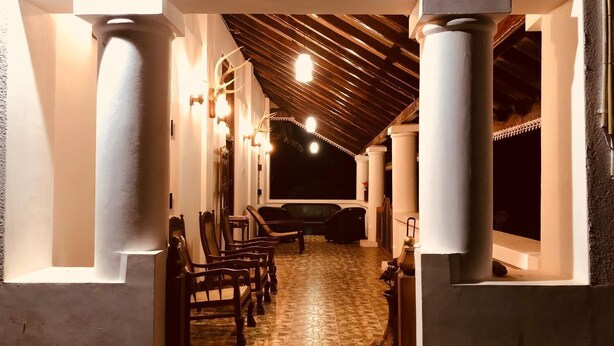 A covered outdoor corridor with wooden benches and columns, leading to a darkened area beyond.