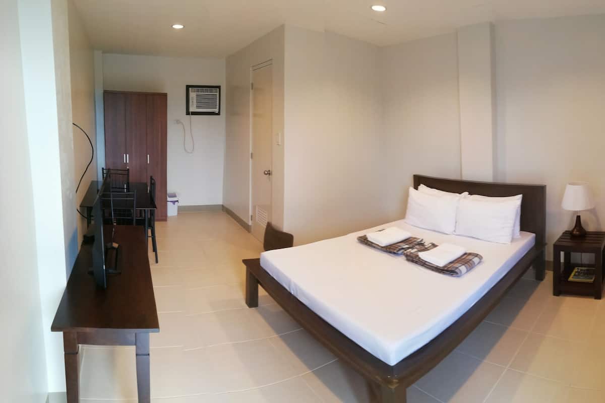 A hotel room with a bed, a desk, a chair, a nightstand with a lamp, and a door leading to another room.