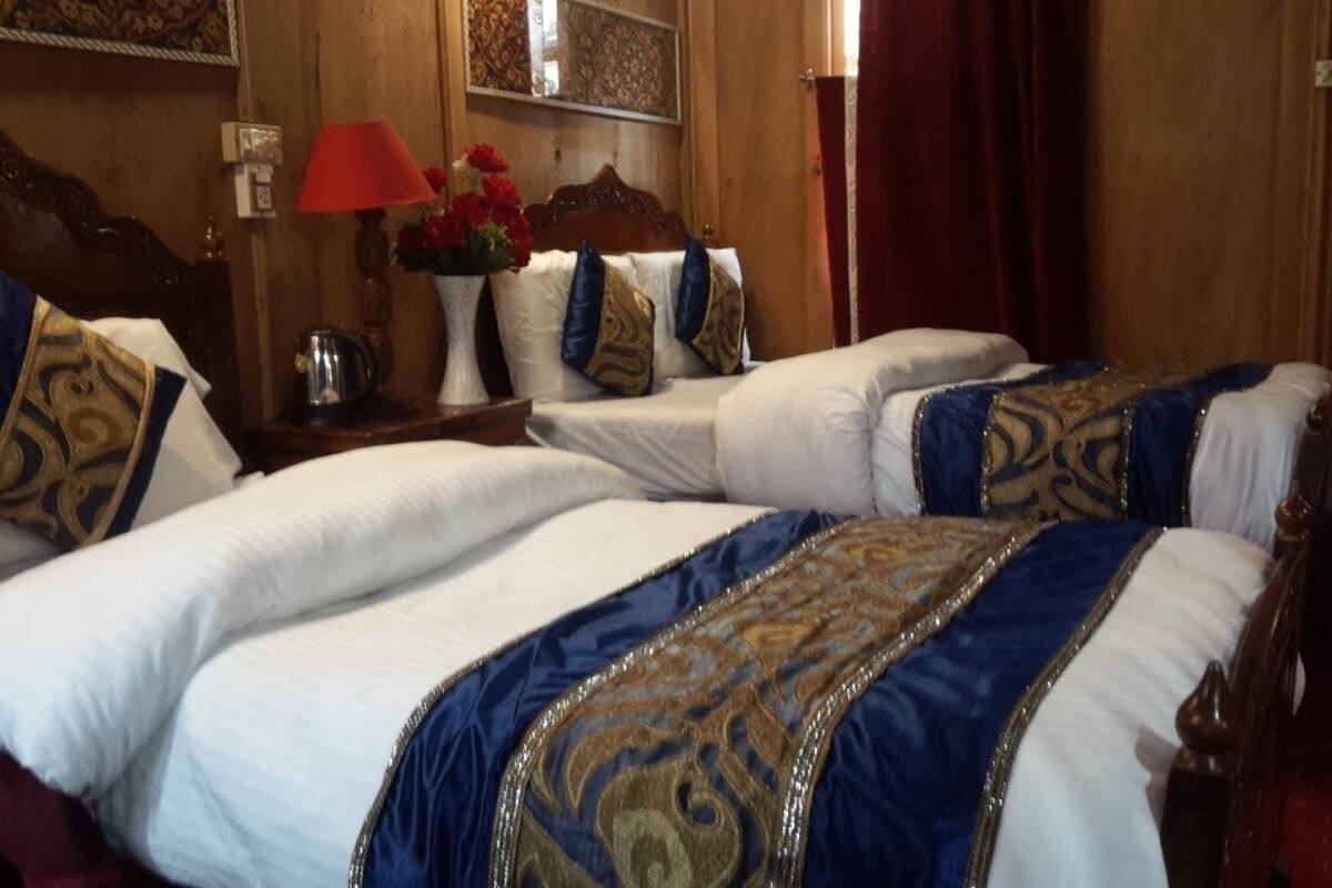 A hotel room with two beds, each with white linens and blue and gold decorative pillows. There is a wooden headboard, a nightstand with a red lamp, and a vase with red flowers.