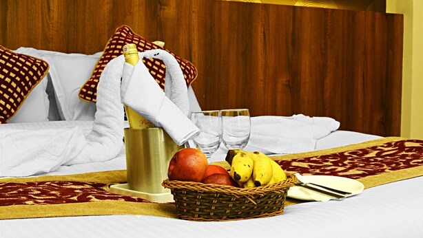 A neatly made bed with a red and gold patterned bedspread, a basket of fruits, a bottle of water, and a pair of glasses on a wooden headboard. - Hotel Royal Suite (Kathmandu)