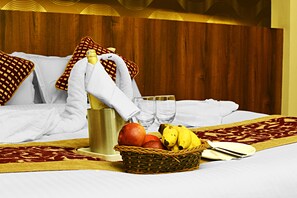 Un lit bien fait, recouvert d’une couvre-lit aux motifs rouges et dorés, un panier de fruits, une bouteille d’eau et une paire de lunettes posées sur une tête de lit en bois.