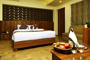 A hotel room with a wooden bed, a wooden nightstand, a wooden table with a fruit basket and wine glasses, and a patterned wall.