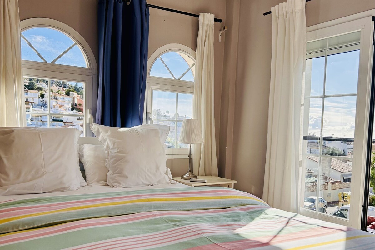 A hotel room with a large bed, two windows with curtains, a nightstand, and a view of the cityscape.