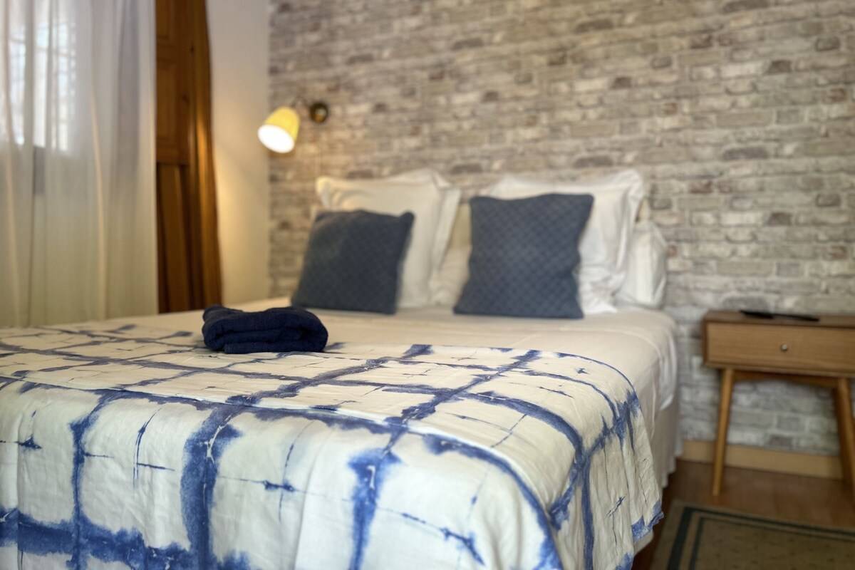 A bedroom with a brick wall, a bed with a blue and white patterned bedspread, two pillows, a bedside table, and a lamp.
