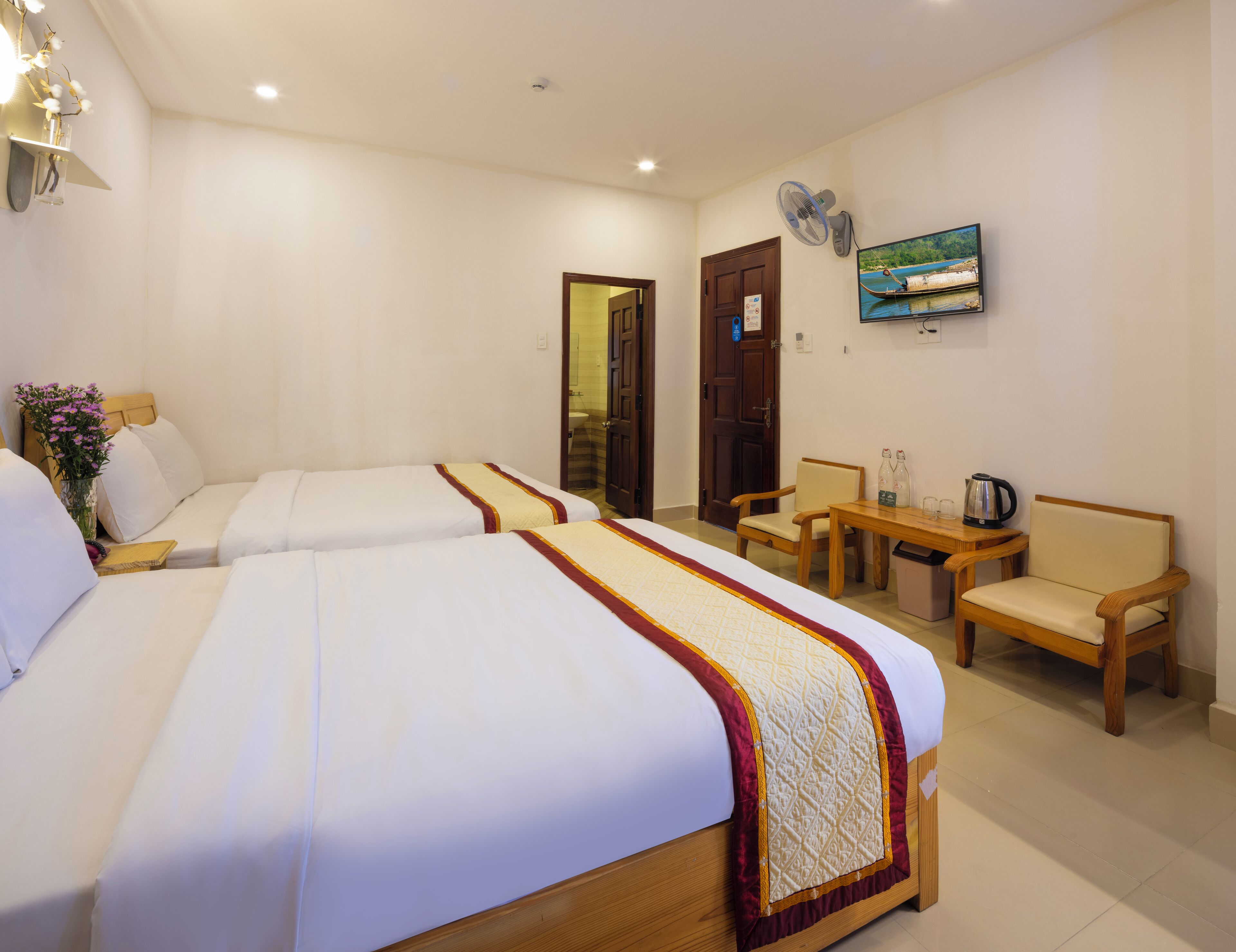 Une chambre d’hôtel avec deux lits, une table en bois, une chaise, une petite table avec une bouilloire et un ventilateur fixé au mur.
