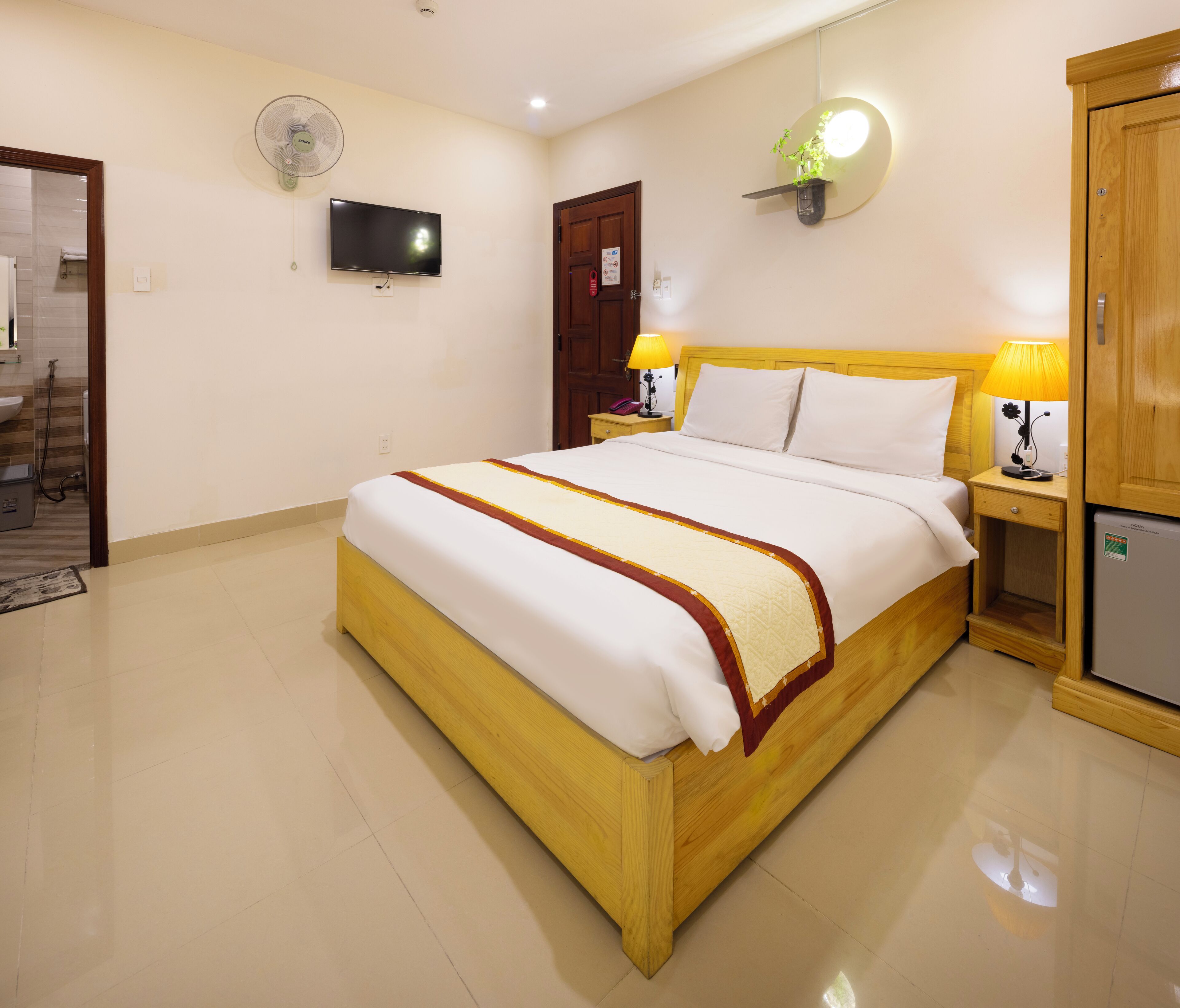 Una habitación de hotel ordenada con cama, lámparas de noche, televisor y un refrigerador pequeño.