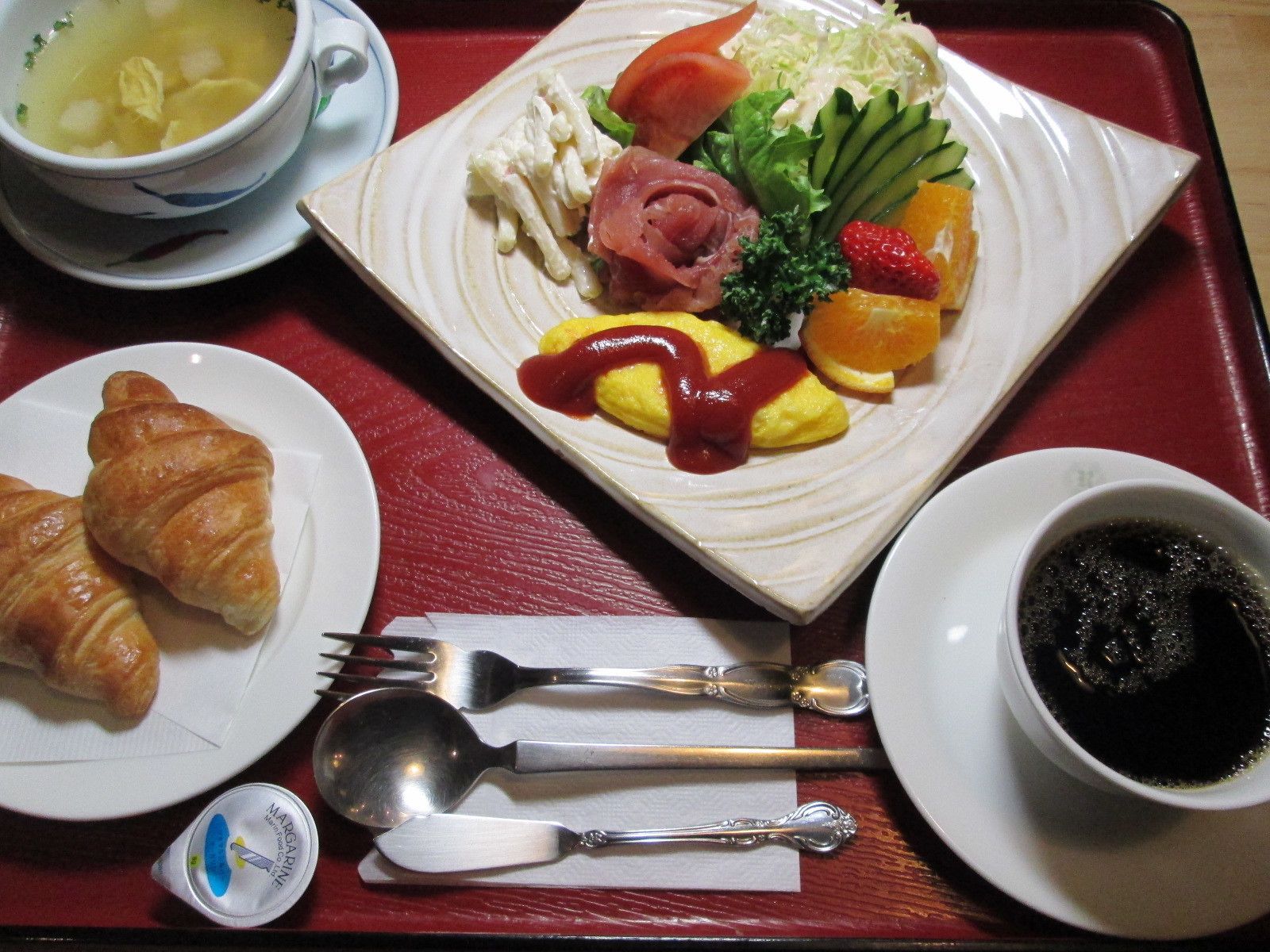 A tray with a plate of food including croissants, a bowl of soup, a plate with ham, vegetables, and a fruit salad, and a cup of coffee.