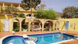 A swimming pool with a stone archway, surrounded by a yellow building with balconies and a mural. - Hotel Mimosa - Hostel (Toubab Dialao)